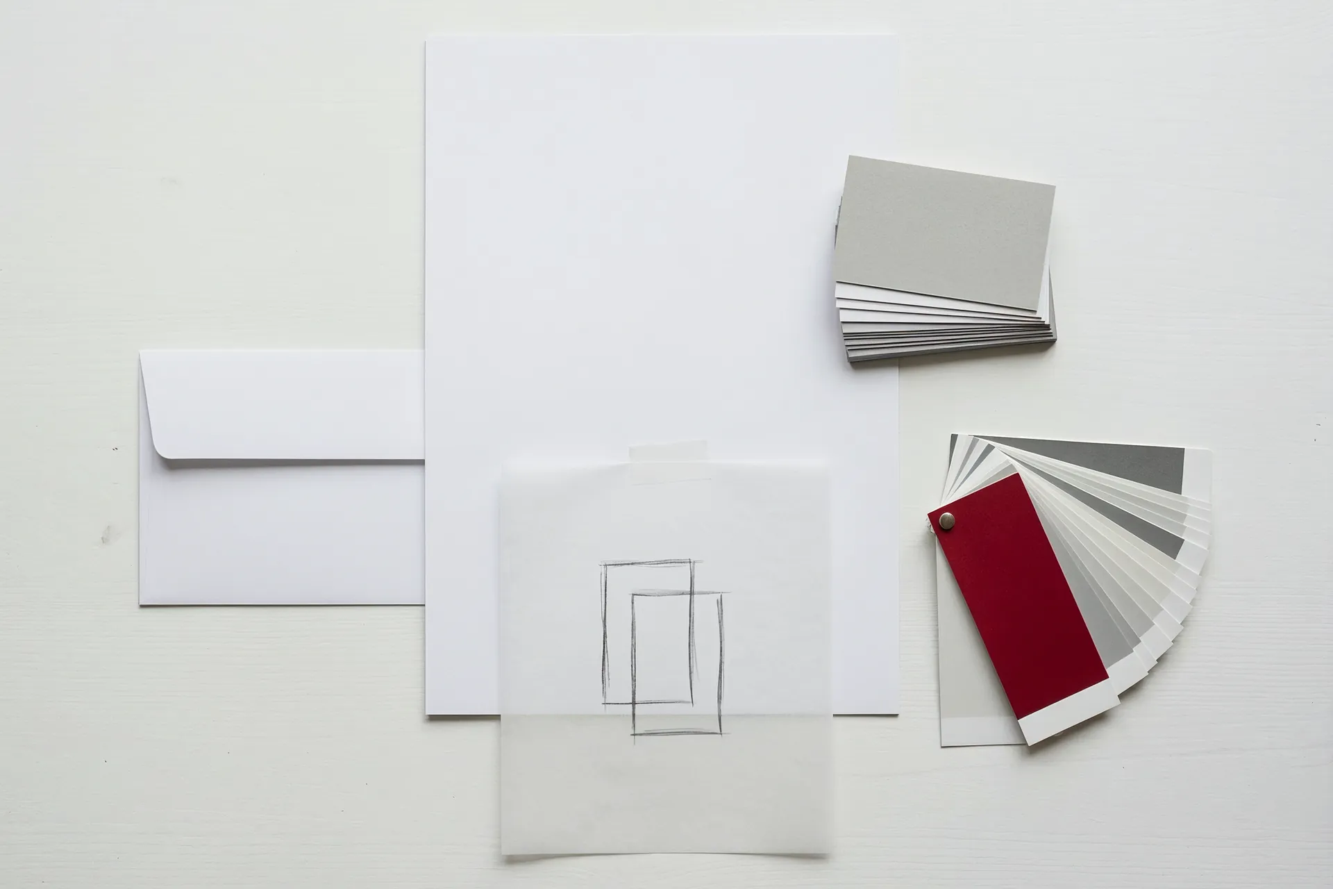 Unbedrucktes Briefpapier, Visitenkarten, ein Umschlag, eine Bleistiftskizze eines abstrakten Zeichens und ein Farbfächer mit einem roten Blatt.